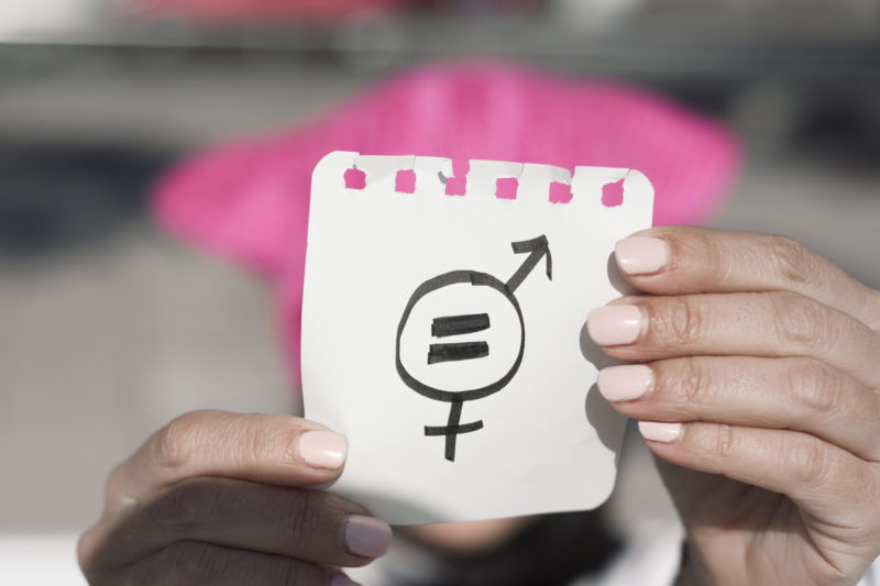 closeup of a young woman outdoors wearing a pink pussycat hat showing a piece of paper in front of her face with a symbol for gender equality drawn in it