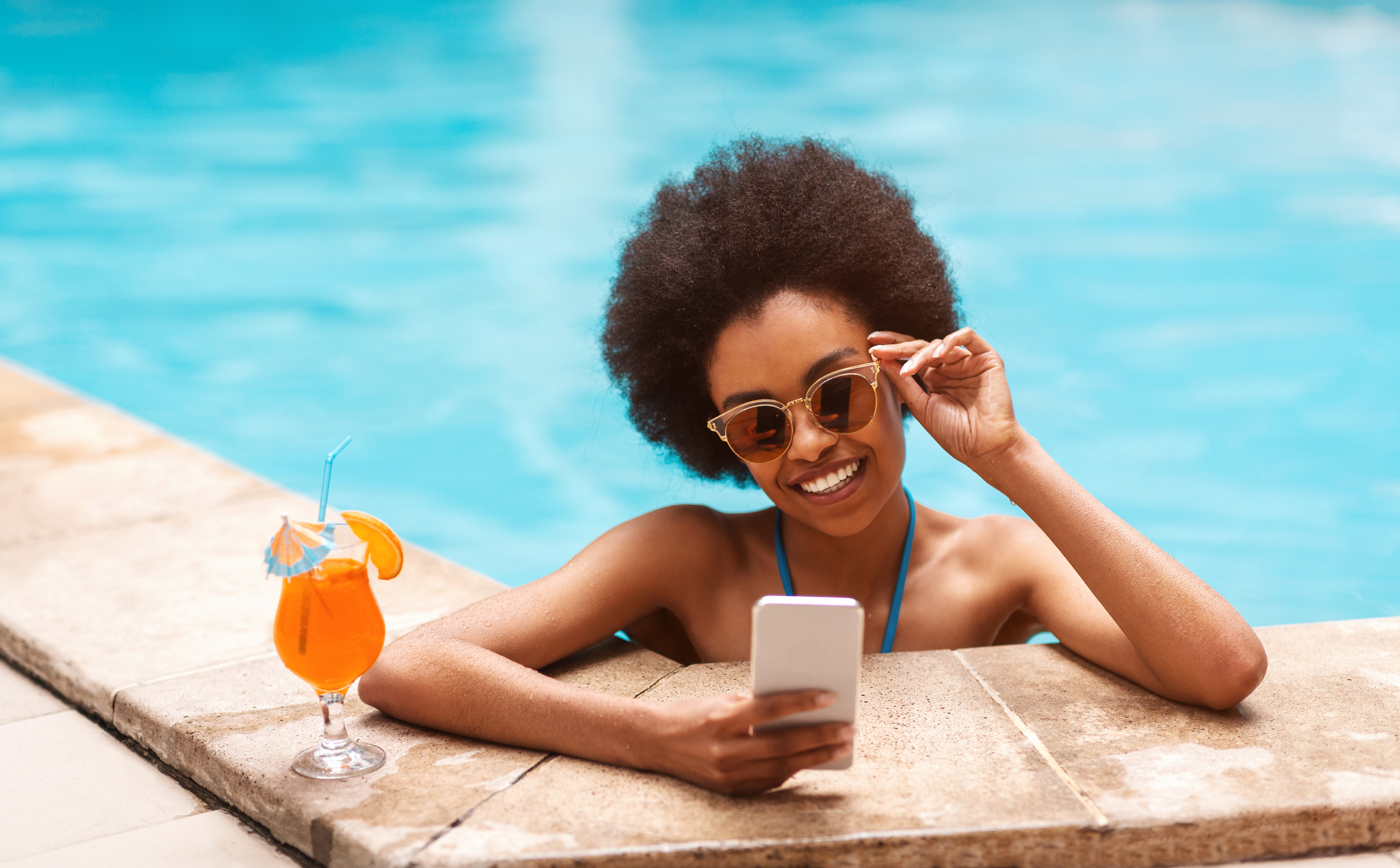 Beautiful black girl with tropical cocktail browsing internet on smartphone at outdoor swimming pool