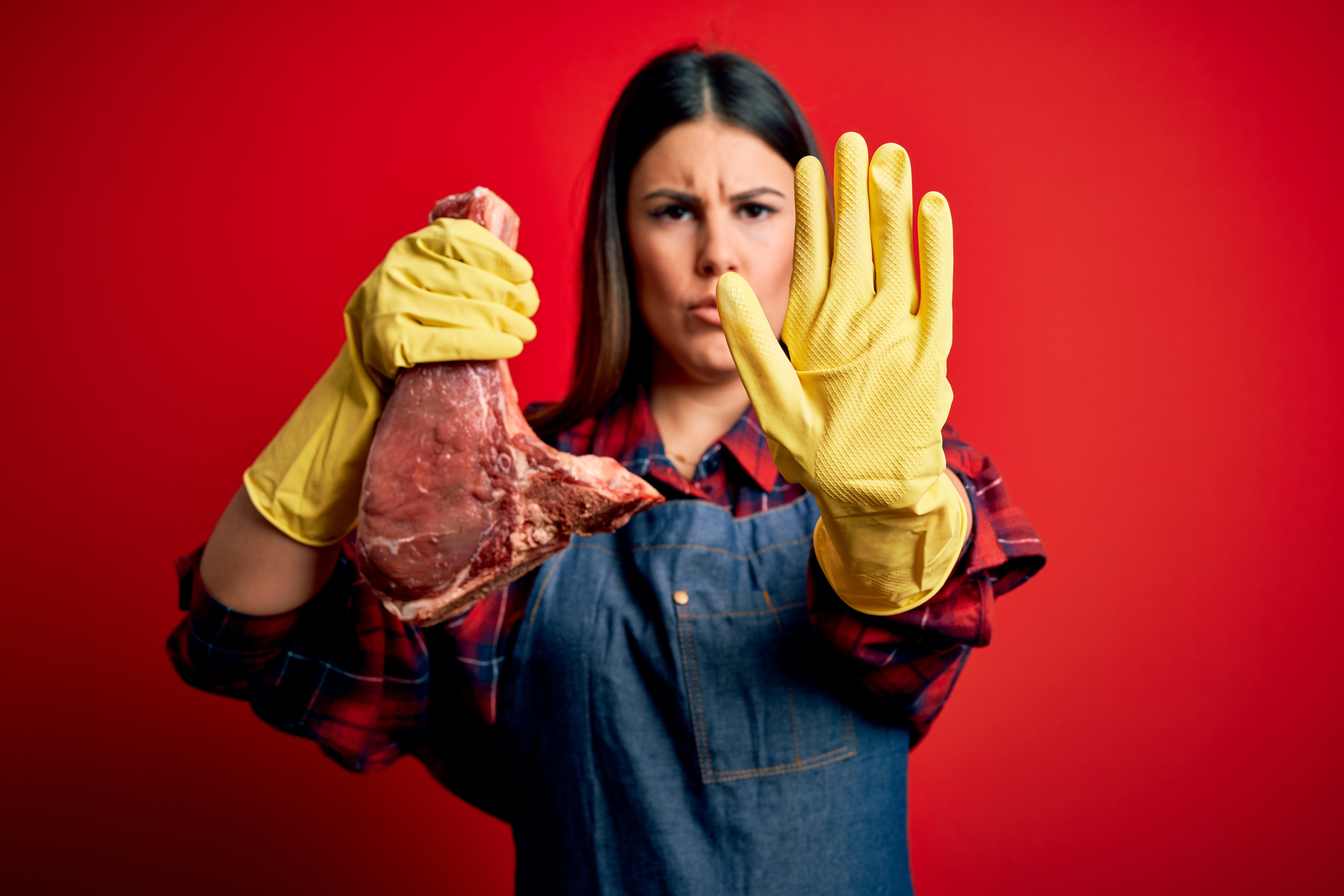 Young butcher woman holding fresh raw beef meat stake over red background with open hand doing stop sign with serious and confident expression, defense gesture