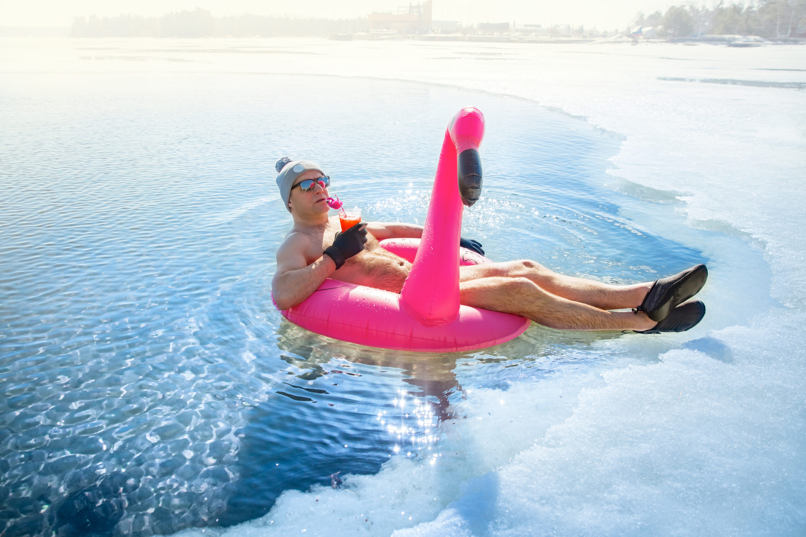 A man swimming in an ice hole in winter in Finland, floating on a pink inflatable flamingo with cocktail in hand. Vacation options, dreaming of summer.