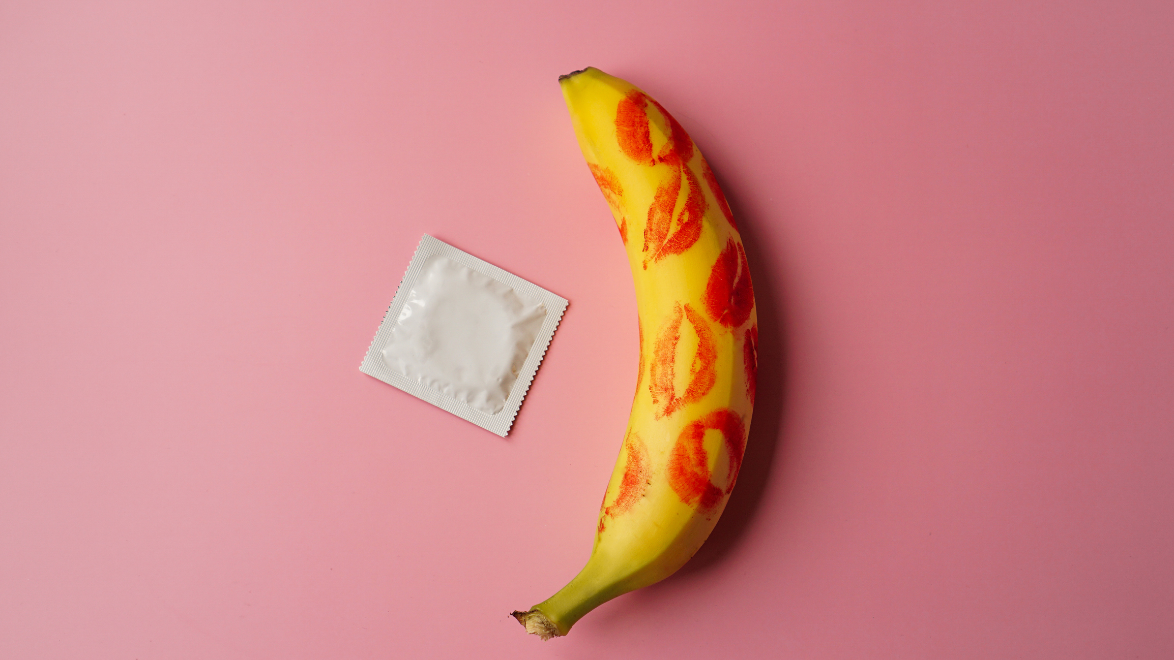 Unpacked condom and red lipstick on a yellow banana on pink studio background. Contraceptives, prevention of venereal disease, preventing pregnancy