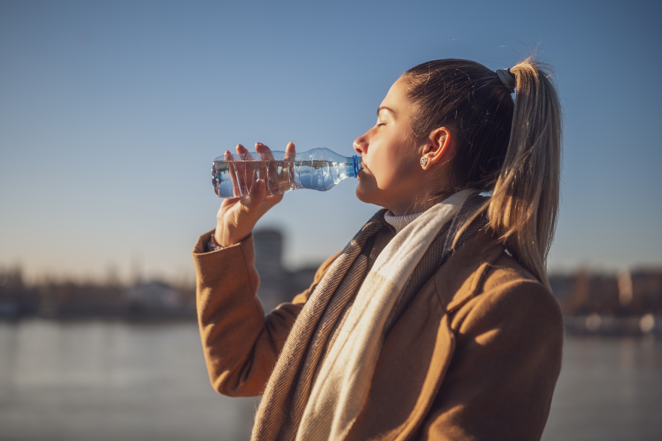 Woman enjoys drinking water while sitting by the river on a sunny winter day. Toned image.