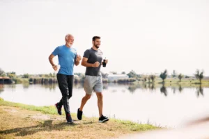 Happy athlete and his senior father jogging by the lake in nature. Copy space.