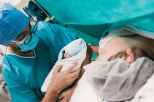 High angle of male obstetrician in medical uniform and glasses demonstrating infant in blanket to mom after C section surgery in hospital