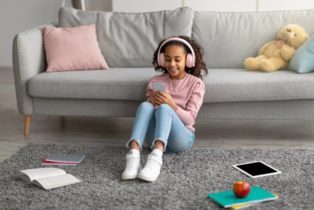 Happy african american girl listening to music in wireless headphones and chatting on her smartphone, taking break from study. Schoolgirl sitting on carpet. leaning on sofa in living room
