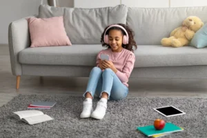 Happy african american girl listening to music in wireless headphones and chatting on her smartphone, taking break from study. Schoolgirl sitting on carpet. leaning on sofa in living room