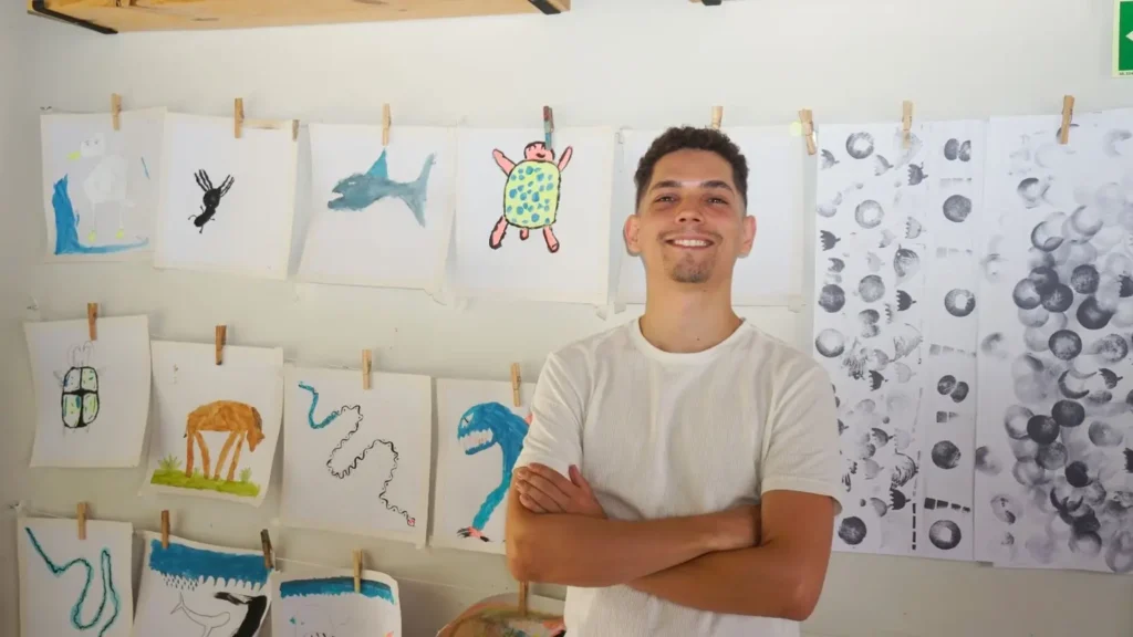 Lucas Liandro posa sorridente em frente às atividades artísticas de alunos com os quais trabalha | Foto: Leandro Braz/O Hoje