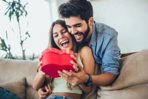 Beautiful young couple is celebrating at home. Handsome man is giving his girlfriend a gift box