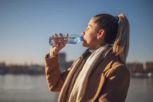 Woman enjoys drinking water while sitting by the river on a sunny winter day. Toned image.