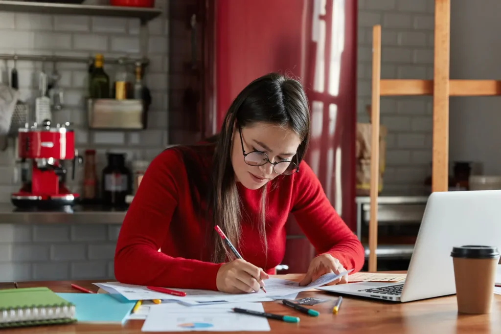 foto horizontal de uma mulher de negocios de aparencia agradavel trabalha com papeis em casa prepara o documento relatorio preenche as informacoes senta se na frente laptop aberto na cozinha