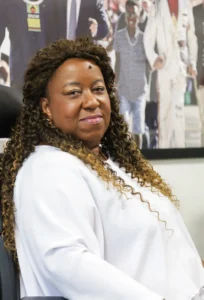 A ministra dos Direitos Humanos e da Cidadania, Macaé Evartisto, é uma mulher megra, de cabelos longos. Na foto, Macaé está de lado, sorri de forma contida, usa uma blusa lisa branca, com mechas do cabelo de cor escura sobre os dois ombros, com as pontas do cabelo mais claras. Ela tem um pinta na testa, usa batom rosa mais claro e sombras mais próximas de um tom amarelado sobre os olhos